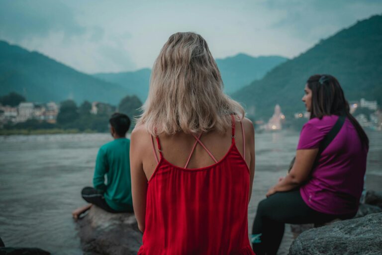 Three people meditate by the Ganges River in Rishikesh, India, with a serene mountain backdrop.